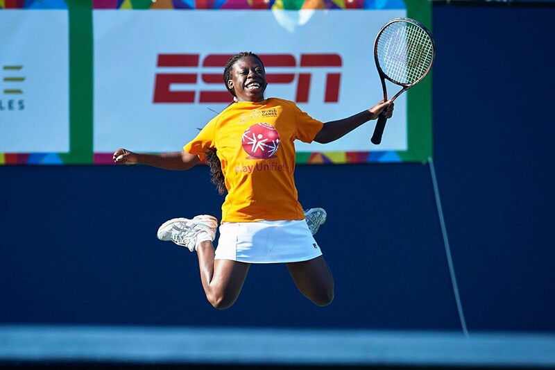 Female tennis athlete jumping up in the air for joy. She's holding her racquet in her left hand, has a white tennis skirt and a yellow Special Olympics t-shirt.