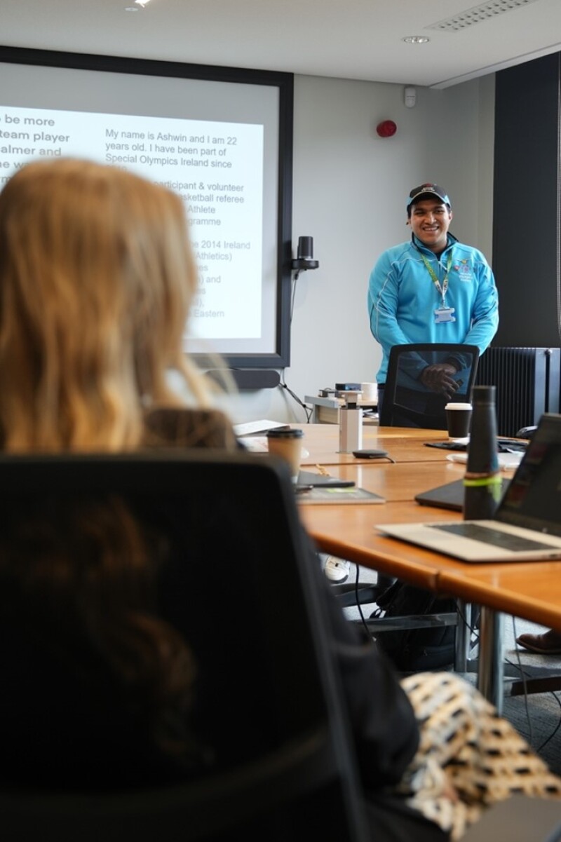 Un hombre con una cremallera azul y un sombrero ofrece una mesa a la gente. Al fondo hay una presentación de diapositivas con información sobre sus logros en las Olimpiadas Especiales, y en primer plano está la parte posterior de la cabeza de una mujer mientras escucha. 
