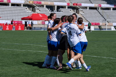 Un grupo de jugadoras de fútbol, vestidas con uniformes blancos y azules, celebran en un círculo de equipo en el campo de un gran estadio, con las gradas prácticamente vacías al fondo.