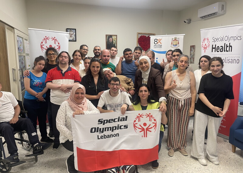 A large group of people holding a flag with the Special Olympics logo smile for a picture.