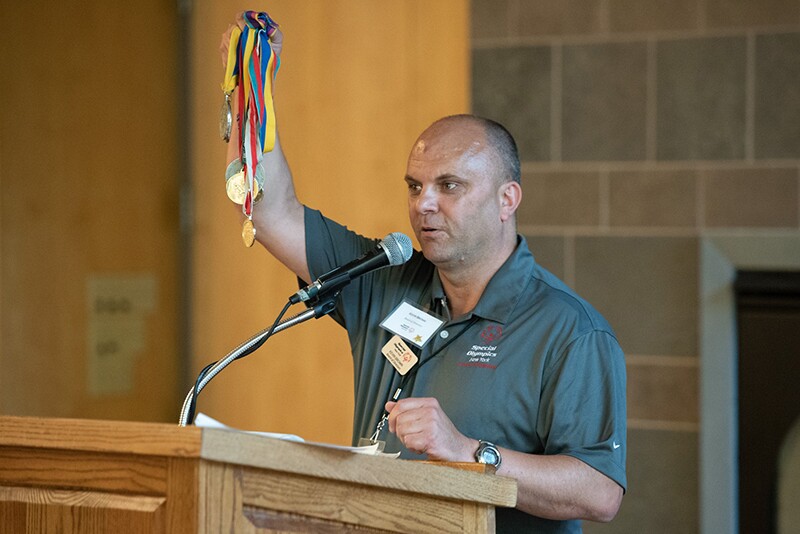 Kevin stands at a podium holding five medals up in the air and stares out to the audience.