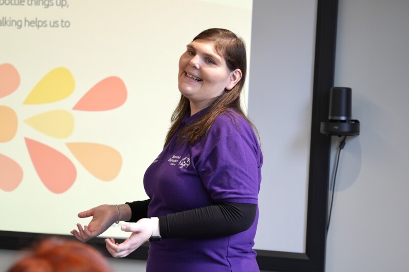 Una mujer que lleva una camiseta morada de Olimpiadas Especiales de Irlanda se pone de pie e inclina la cabeza hacia atrás mientras presenta una diapositiva sobre salud mental. 