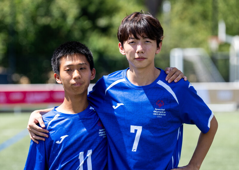 Two football teammates wearing blue Special Olympics China jerseys stand side by side on a football field with their arms around each other's shoulders. The player on the left wears number 11, while the player on the right wears number 7. Trees and a goalpost are visible in the blurred background.