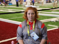 Emileigh Marsh sits in a wheelchair on an indoor turf competition field, wearing a gray and red Texas team shirt and a yellow credential lanyard. Bocce courts and event participants are visible in the background.