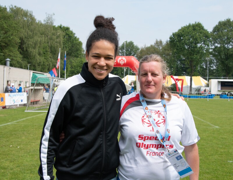 Two women stand on a football pitch facing the camera and smiling. There is a football/soccer net and an inflatable arch in the background.