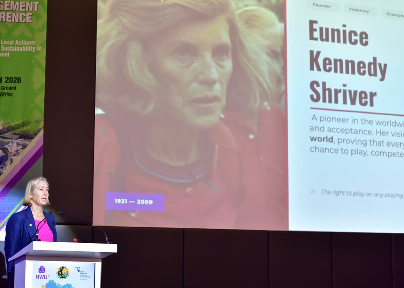 A woman stands behind a podium while she speaks during a presentation. A large screen is behind her showing a PowerPoint presentation.