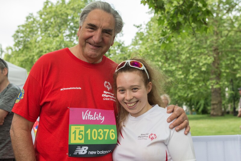 A man in a red Special Olympics t-shirt smiles to the camera with his arm around a young woman in a white Special Olympics t-shirt.