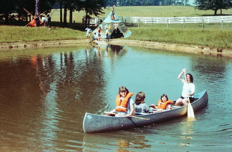 Niños y monitores participan en actividades al aire libre en el Camp Shriver de la década de 1960, con una canoa en un lago y tiendas de campaña al fondo.