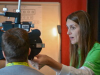A health volunteer in a green Special Olympics Health shirt conducts a vision screening. A Special Olympics athlete with this back turned to the camera looks through screening equipment. In the distance a LEA eye chart which uses symbols instead of letters is visable. 