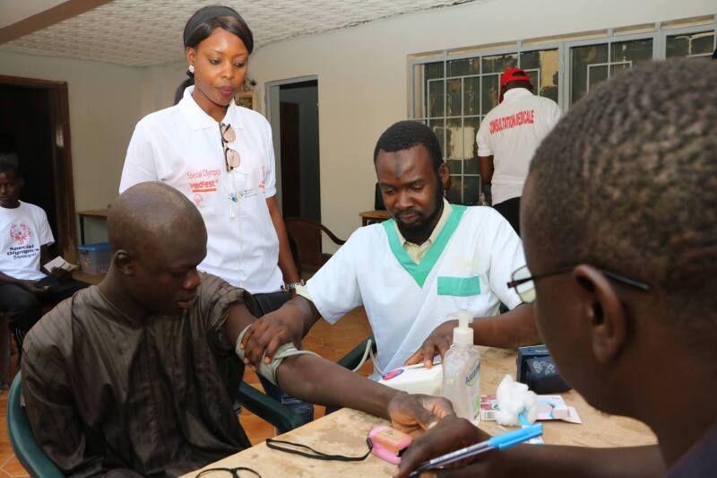 Athlete receiving a blood pressure check by a health volunteer; another volunteer is observing and another is recording results.