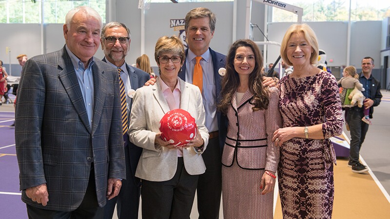 A group of men and women in professional clothing hold a red soccer ball in a gymnasium.