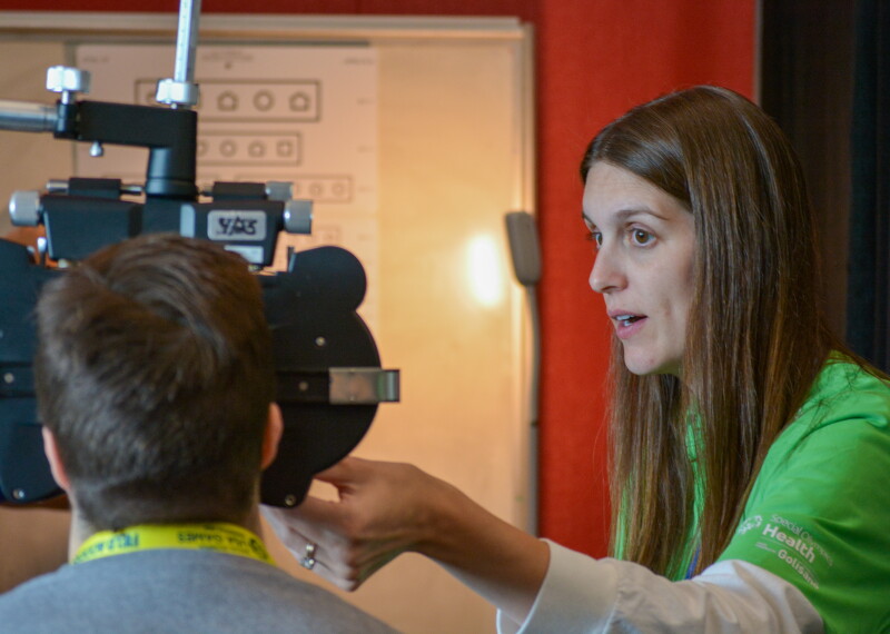 A health volunteer in a green Special Olympics Health shirt conducts a vision screening. A Special Olympics athlete with this back turned to the camera looks through screening equipment. In the distance a LEA eye chart which uses symbols instead of letters is visable. 