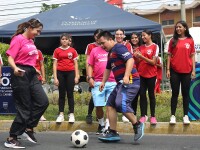 A young boy wearing a Barcelona football kit smiles while playing football with a cheerful woman in El Salvador, as girls cheer in the background.