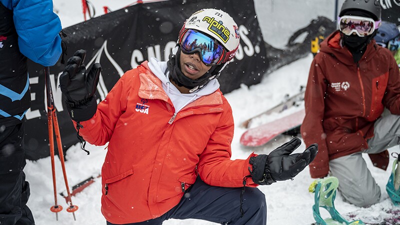 Latrice in and orange ski jacket on one kneed posing for the camera on the slopes.