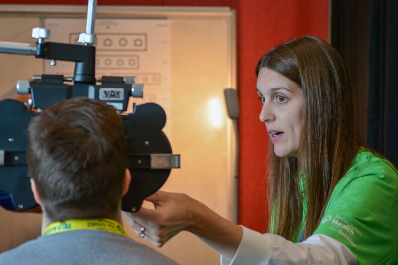 A health volunteer in a green Special Olympics Health shirt conducts a vision screening. A Special Olympics athlete with this back turned to the camera looks through screening equipment. In the distance a LEA eye chart which uses symbols instead of letters is visable. 