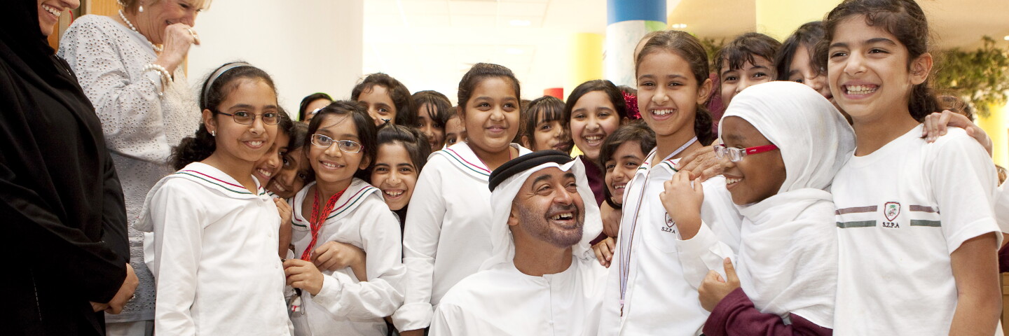 His Highness Sheikh Mohamed Bin Zayed Al Nahyan, President of the United Arab Emirates sitting down and interacting with daughter on his right and her classmates stand to their left and right.