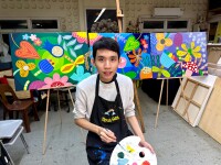 A young man stands in front of 3 paintings holding paint and a paint brush.