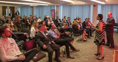 A female and male speaker stand at the top of the room addressing a seated audience.