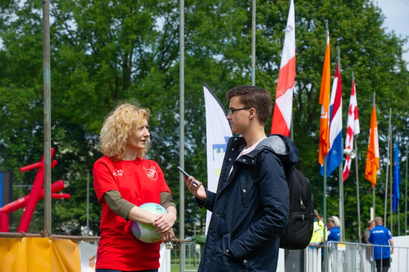 Woman in a red t-shirt holding a colourful ball speaks to a man holding a phone with flags in the background. 