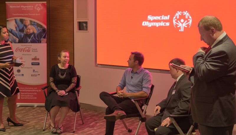 Three people sit in front of a screen branded with the Special Olympics logo. A female and male stand at either side of them.