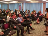 A female and male speaker stand at the top of the room addressing a seated audience.