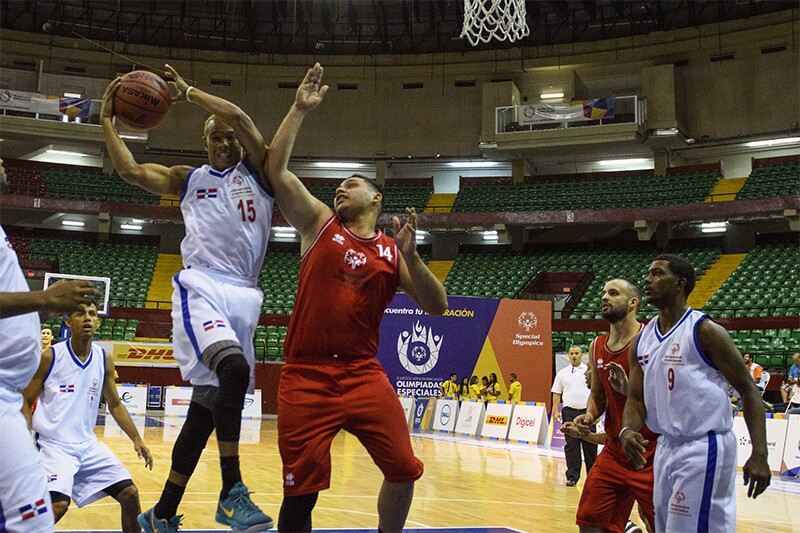 Six player can be seen on the basketball court. One athlete is jumping for a dunk and a player for the opposing team is jumping to defend.