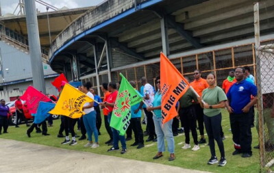 A group of people stands outdoors along a walkway beside a stadium or large sports venue. Several participants hold brightly colored flags, including orange, green, yellow, blue, and red flags. The orange flag in the foreground displays the text “THE BIG MIX.”