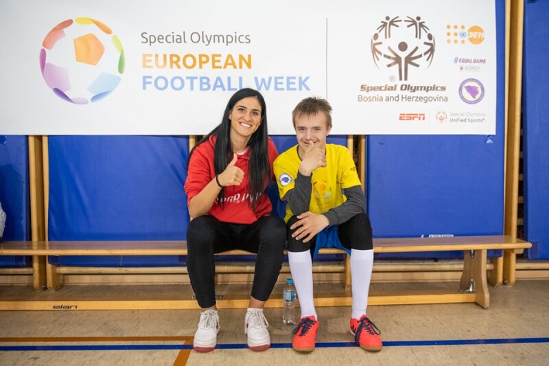 A woman and young man sit on a bench smiling at the camera with their thumbs up in the air. Behind them is a Special Olympics European Football Week banner.