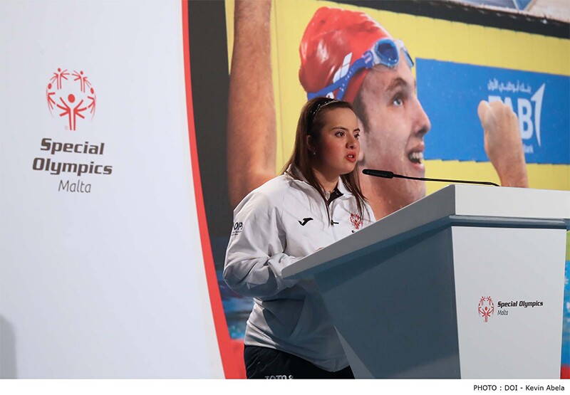 A young woman speaks into a microphone on a podium with the Special Olympics Malta logo on the podium and in the background.