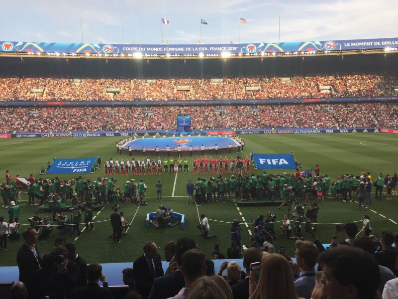 View of a football pitch in a crowded stadium with teams lined up on the football pitch. A FIFA flag and a large flag of the FIFA logo are being held up by people on the football pitch.