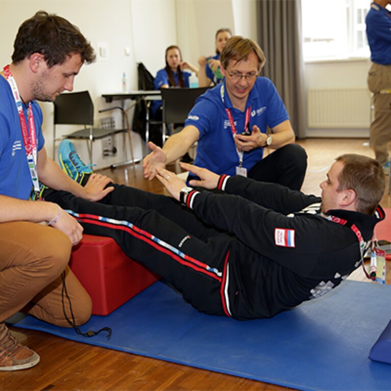 athlete laying on his back with his legs elevated reaching to touch his toes; two volunteers observe and encourage him.