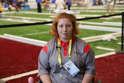Emileigh Marsh sits in a wheelchair on an indoor turf competition field, wearing a gray and red Texas team shirt and a yellow credential lanyard. Bocce courts and event participants are visible in the background.