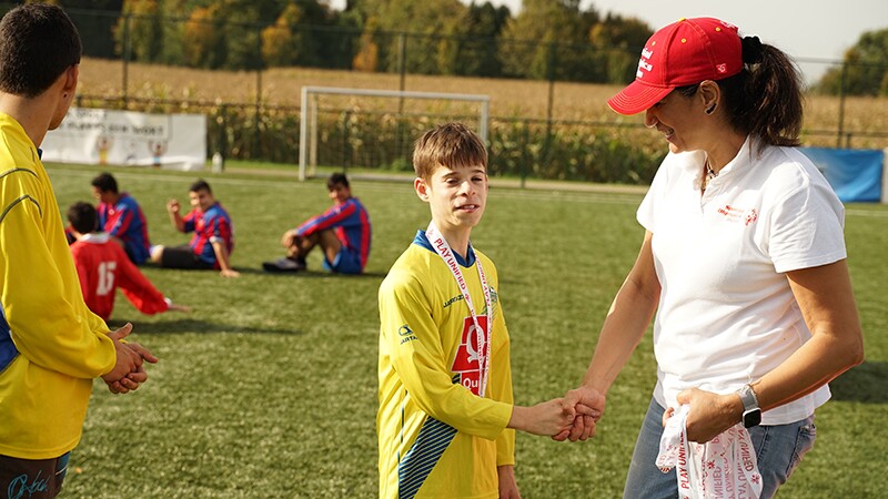 A young man in a yellow football jersey shakes hands with a woman in a white t-shirt and red baseball cap. They are standing on a football pitch and there are other footballers in the background.