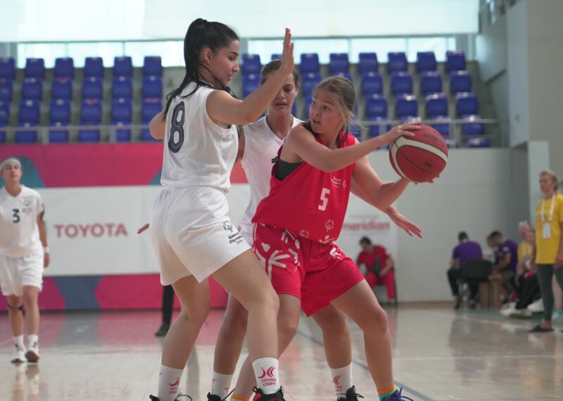 A group of three young girls playing basketball. Two girls wearing white jersey man-marking the third one who is holding the basketball and wearing the red uniform of the opposite team.