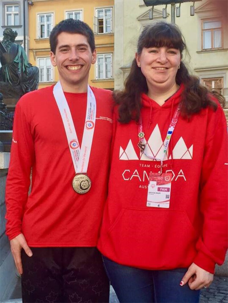 Matthew and Crystal in red shirts, Mathew is wearing a medal around his neck, their arms are around one another and they both are smiling.