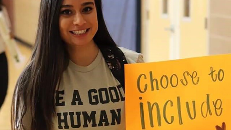 Savyna smiling wearing a shirt that reads, "Be a good human" and holding a sign that reads, "Choose to Include." 