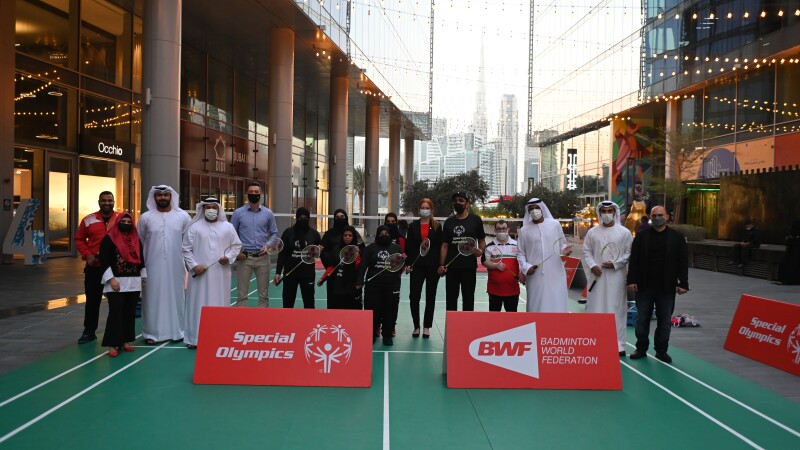 A group of 16 representatives from the various organizations standing outdoors on an Airbadminton court, with organization logos in front of them and the Burj Khalifa in the distant background. 