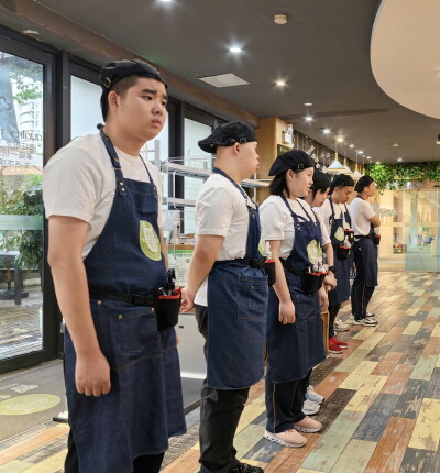 A line of trainees in matching white shirts, dark aprons, and black caps stand side by side inside a brightly lit restaurant or café. They are positioned along a wooden floor near a service counter, with tools or cleaning supplies attached to their waists. Large windows line the left side, and the interior features warm lighting, hanging fixtures, and decorative greenery in the background.