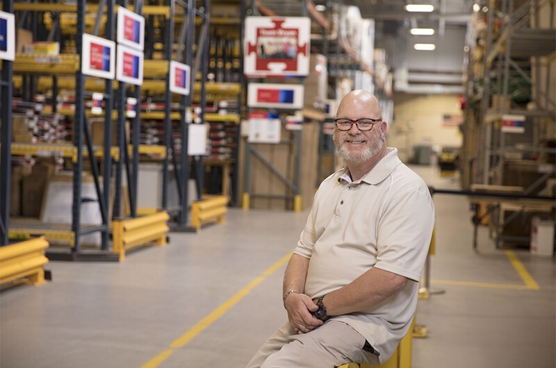 Tommy Fields at work in the Bank of America Support Services marketing fulfillment center. Tom is sitting on a railing in a warehouse facility.