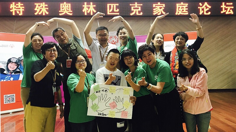 Attendees of the Special Olympics Family Forum in Xi'An, China are in a group, six males and females in the front row and six in the back row, the back row is making a heart shape with the their hands and arms above their head with the person next to them. A young man in the front is holding a poster with notes and a drawing of two hands inside a heart.