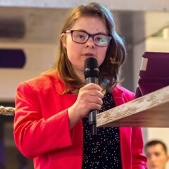 Lize Weerdenburg in a red suite jacket and black shirt with white spots standing behind a podium speaking.
