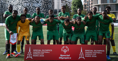 Un equipo de fútbol posa para una foto. Llevan uniformes verdes y están de pie detrás de un cartel rojo que dice "Copa Mundial de Fútbol Unificado de las Olimpiadas Especiales París 2026".