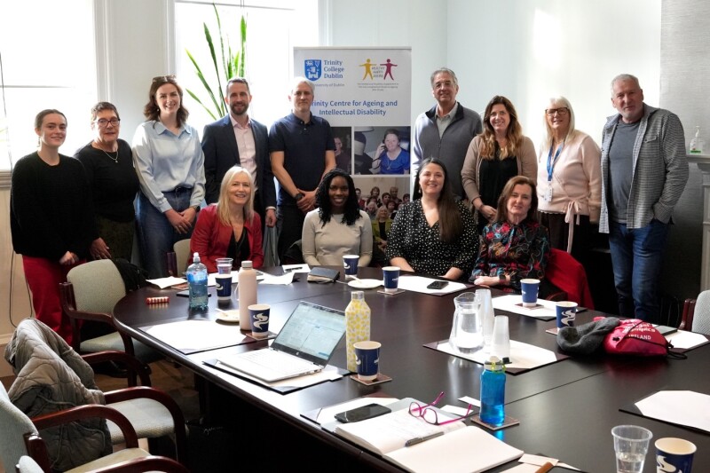 Un grupo de 13 personas posan sonriendo juntas en una sala de reuniones detrás de una gran mesa de conferencias cubierta de cuadernos, papeles, botellas de agua, portátiles y otros objetos. Varias personas están sentadas a la mesa y otras están de pie detrás de ellas. Una pancarta al fondo muestra el Centro Trinity para el Envejecimiento y la Discapacidad Intelectual con su logotipo y una fotografía.