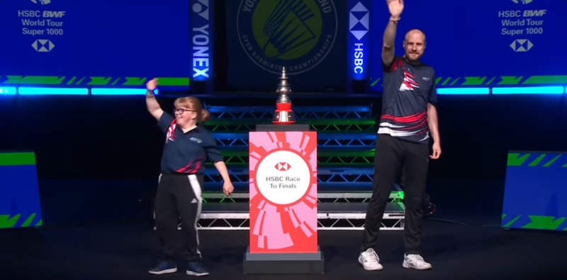 Dee y Clint están parados a ambos lados de un gran escenario rojo sosteniendo el trofeo oficial y agitándolo ante la audiencia. 