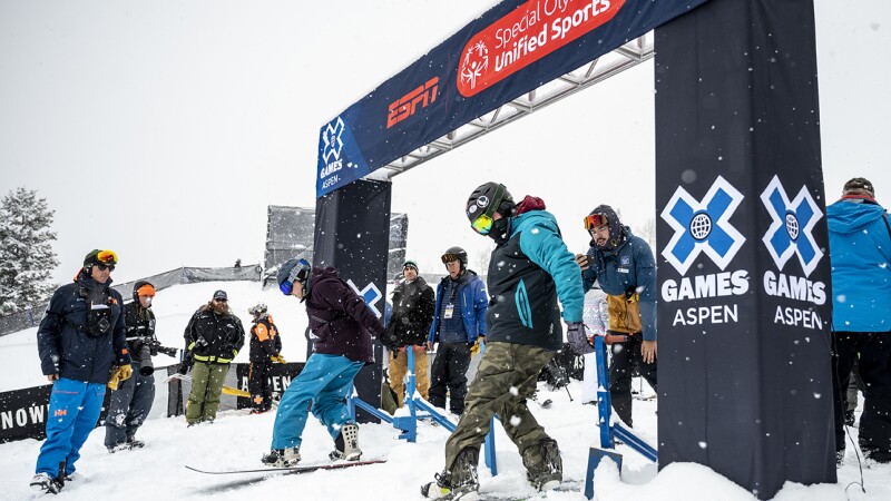 Two people are on snowboards under the starting sign waiting for their race to start. Other people are standing and watching them.
