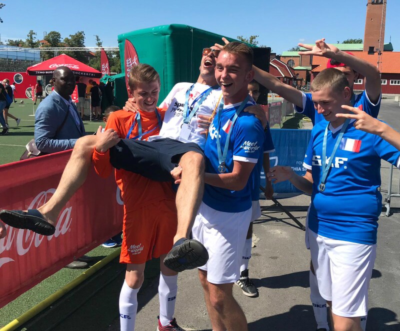 A group of boys in football kits carry a man in a white t-shirt and sun glasses. They are all smiling and some of the boys have their hands in the air.