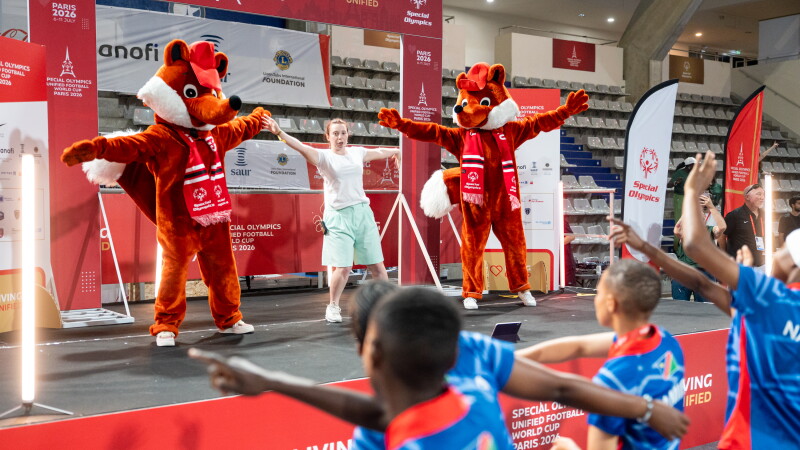 Young woman dancing on stage with two mascots and players join in the dance.  