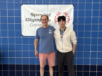 The photo shows an adult man standing next to a male high school student. They are in front of a blue wall with a Special Olympics Connecticut banner hanging on it. 