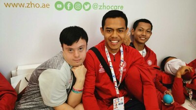 Four young men sitting in a group, two have on long sleep red Special Olympics Abu Dhabi 2019 pull overs. 