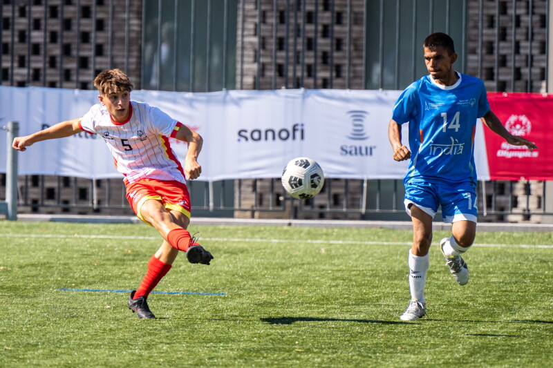 Un jugador contrario con uniforme blanco y rojo patea el balón mientras un jugador con uniforme azul corre a su lado para detener el juego durante un partido de fútbol al aire libre. Pancartas de los patrocinadores se alinean en la valla detrás del campo de césped artificial.